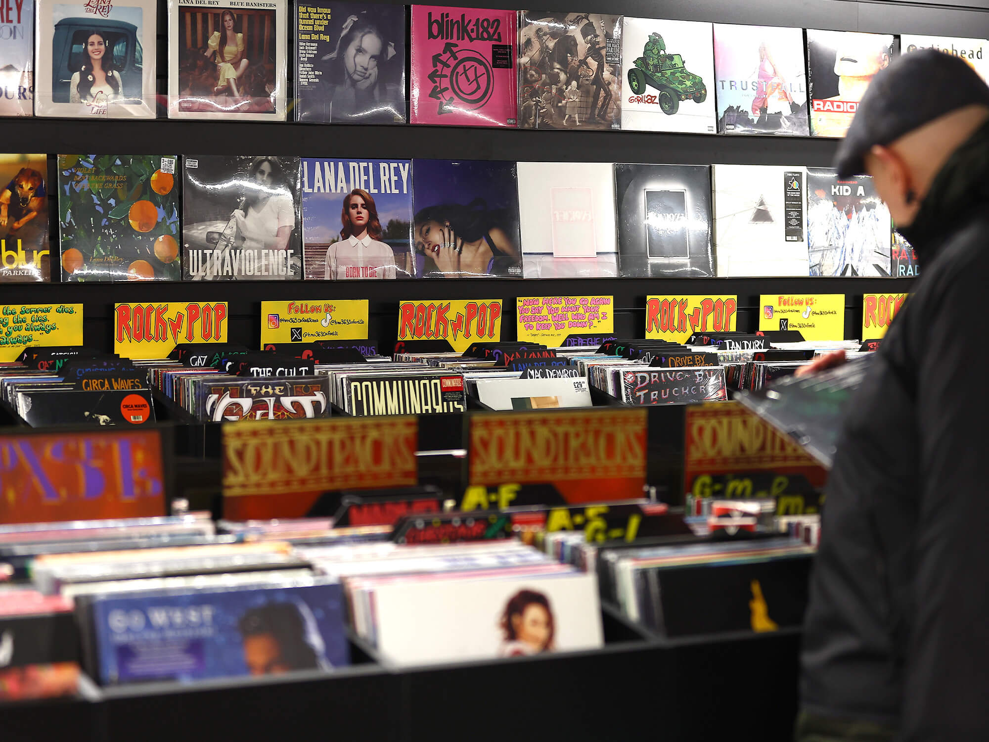 Vinyl records displayed on the wall in HMV. There are also large boxes filled with vinyls, which are sorted under labels such as "rock" and "pop". A person stands to the right of the photo, holding one of the records.