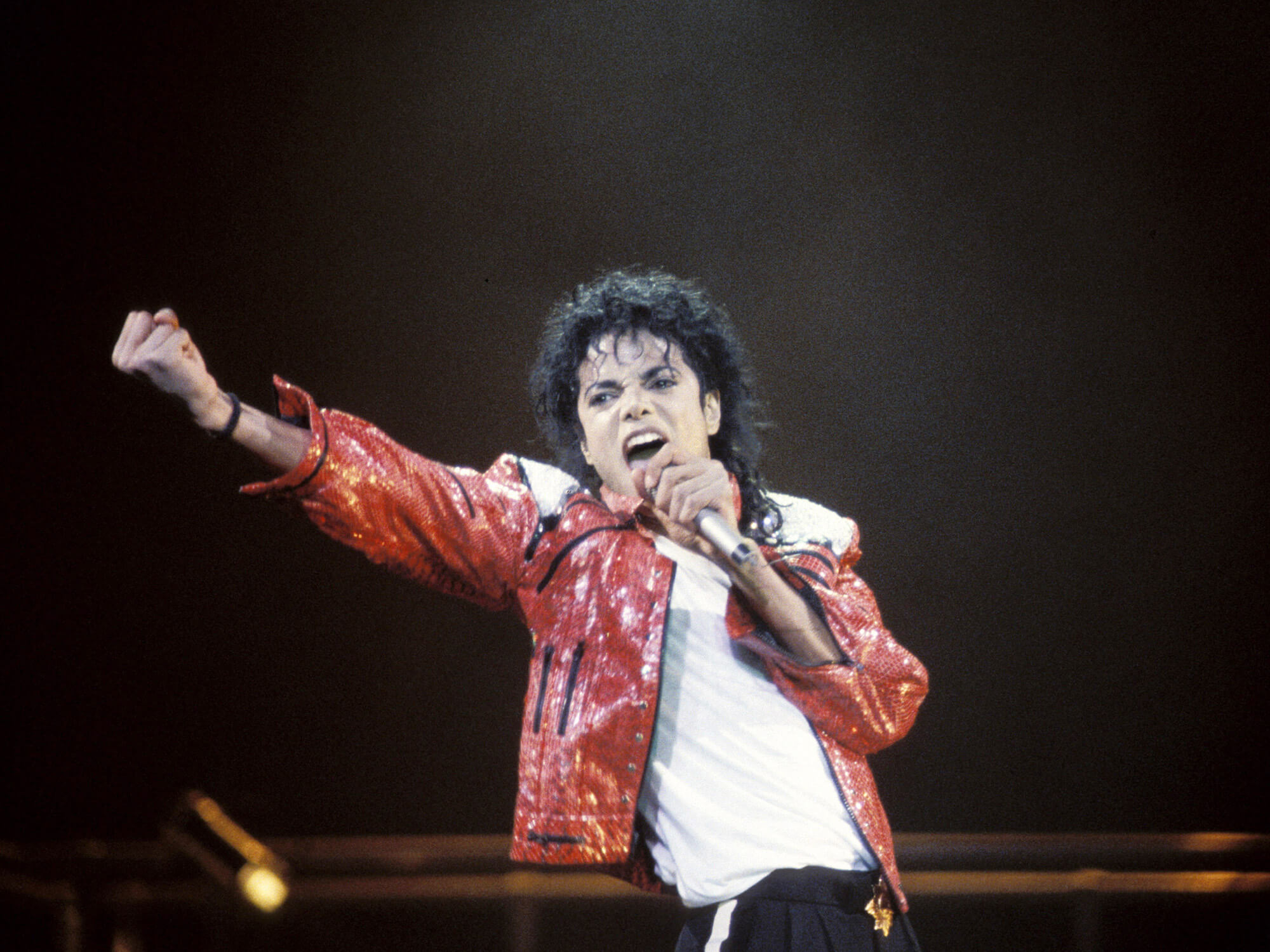 Michael Jackson in his famous bright red jacket. He is on stage, holding a mic in one hand and his other hand up in the air clenched in a fist as he is singing.