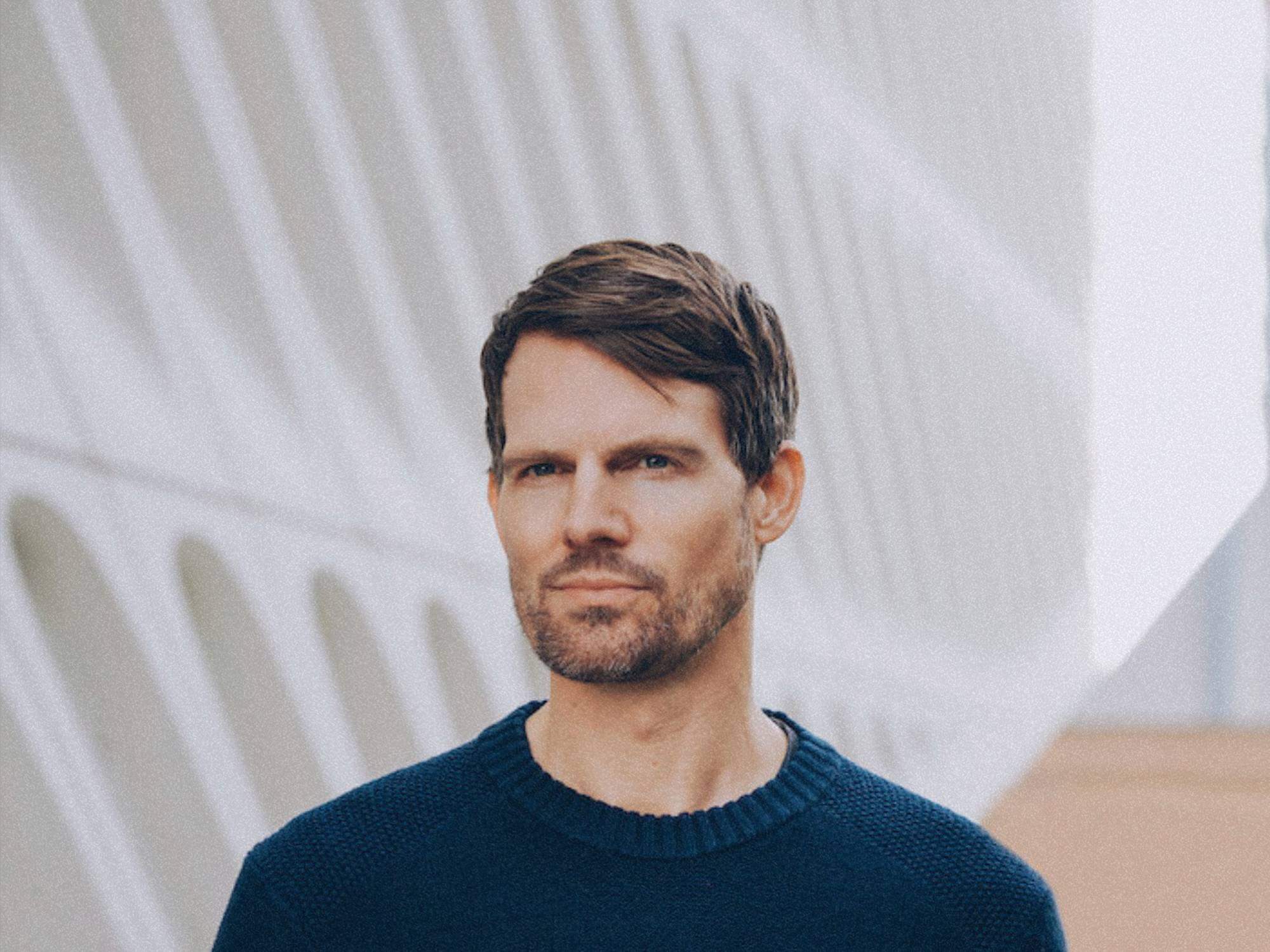 Tycho stood in front of a white background. He's wearing a plain t-shirt and is looking at the camera.